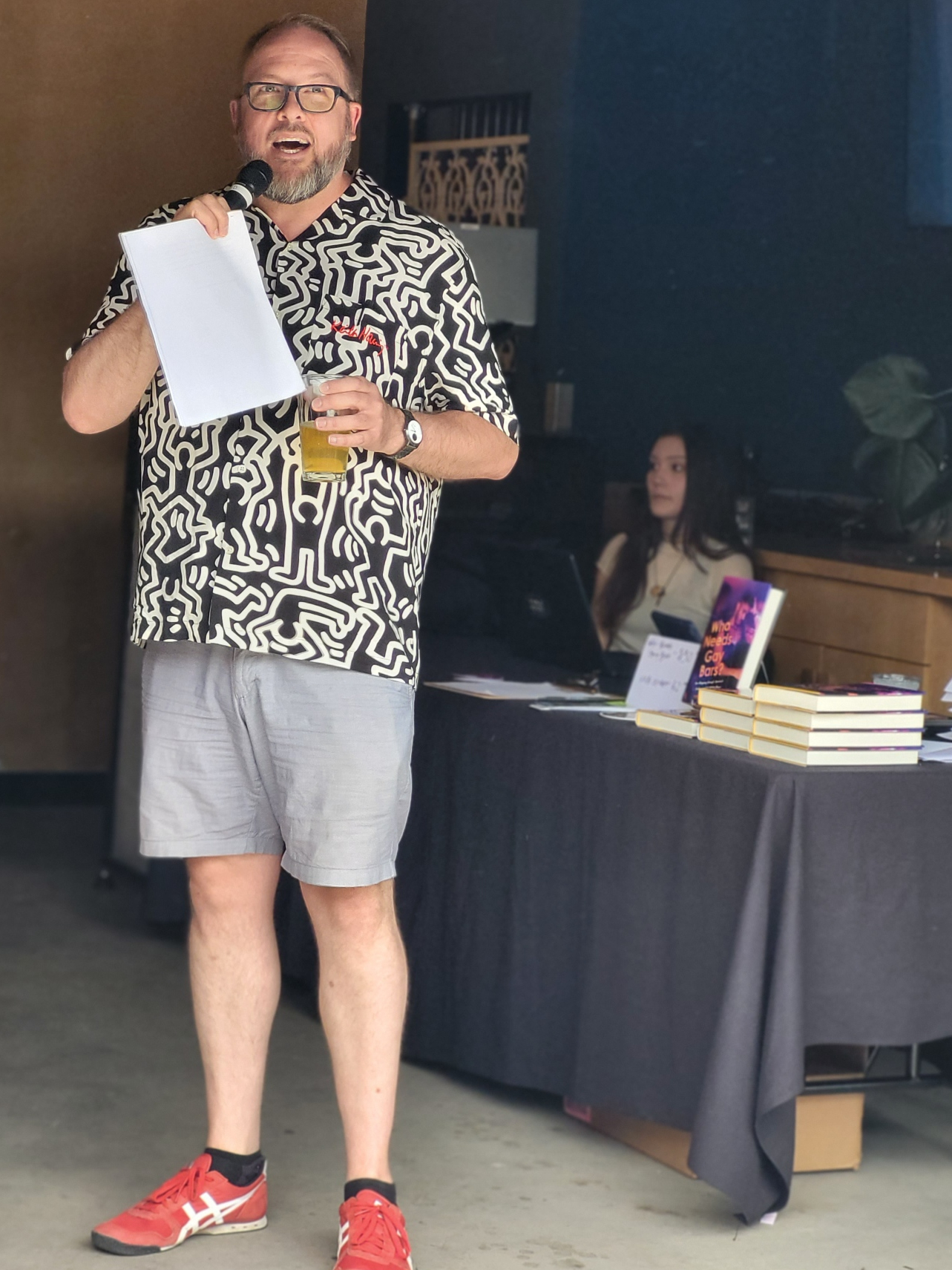 White man in Keith Haring black and white shirt and red sneakers standing with a beer in his hand and talking